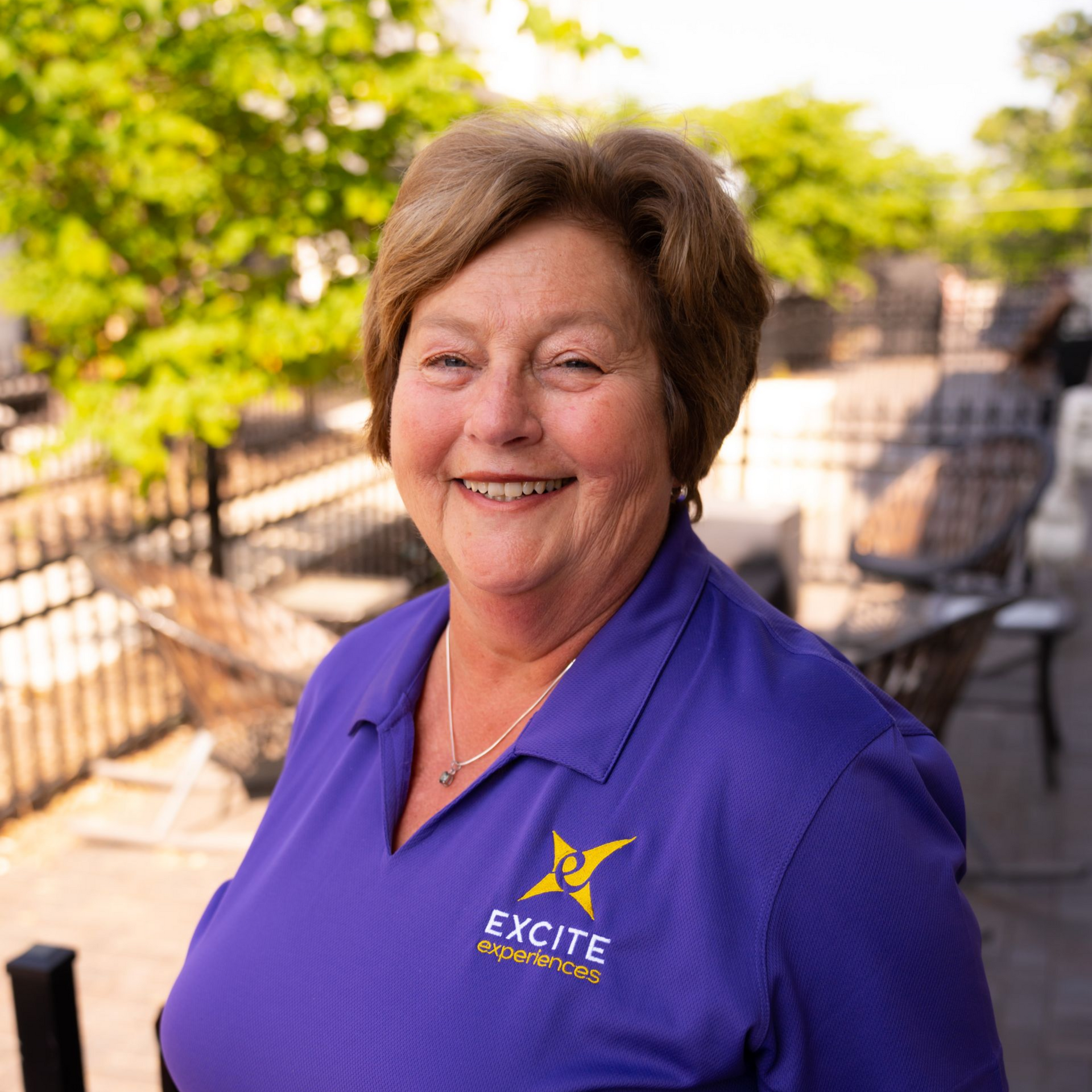 Woman in purple shirt smiles outdoors near greenery, with