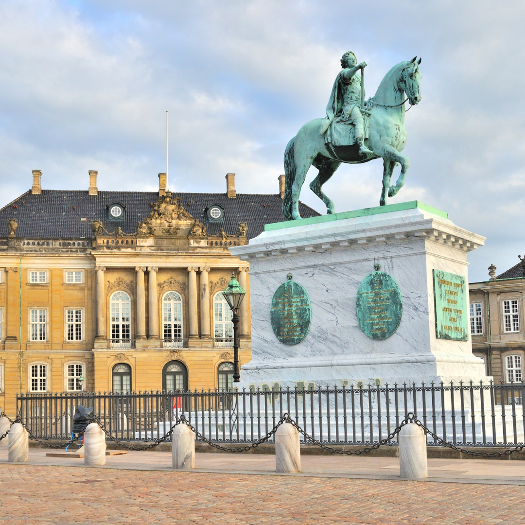 Equestrian statue of a man atop a white marble pedestal in front of a yellow palace in Copenhagen.