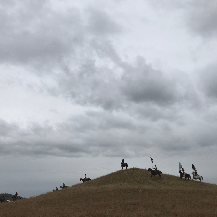 Horses with riders on a grassy hill under a cloudy sky.