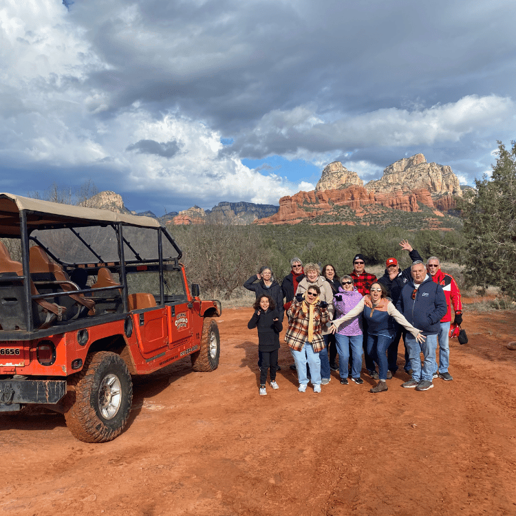 Group poses near red jeep and red rock formations in Sedona, Arizona under cloudy sky.