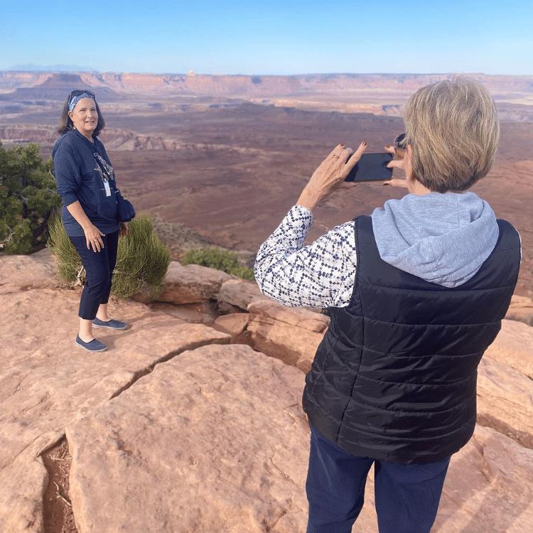 Woman taking photo of another woman on a red rock plateau overlooking a desert vista.