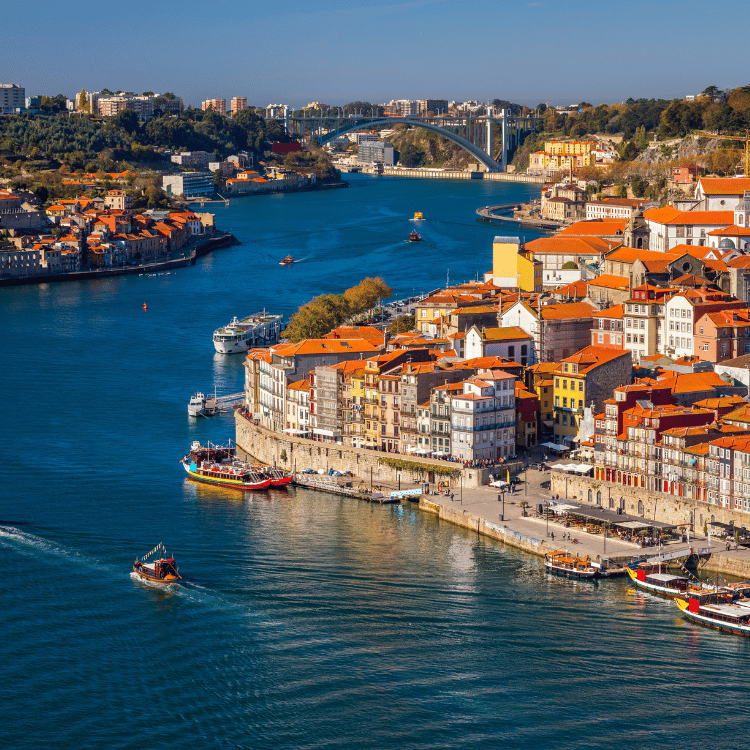 Waterfront cityscape with orange rooftops, blue river, boats, and a bridge.