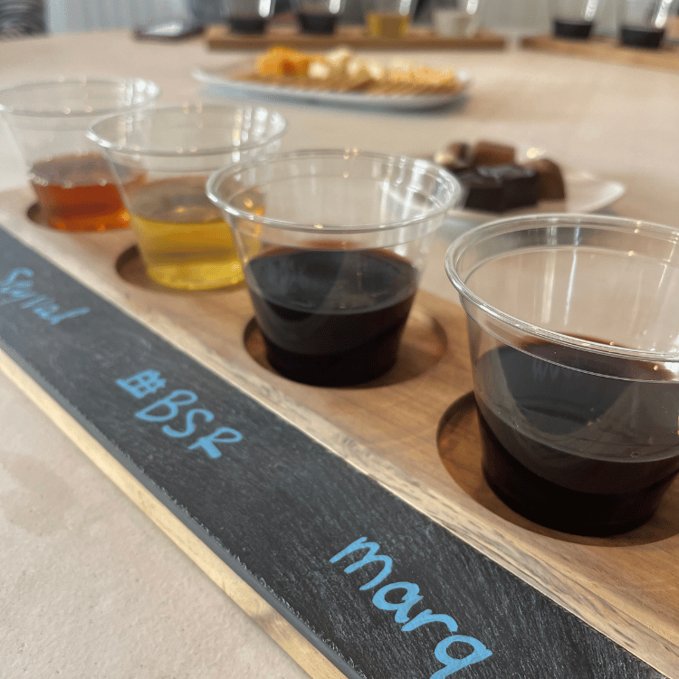 A flight of wine in clear cups on a wooden tray, with snacks in the background.