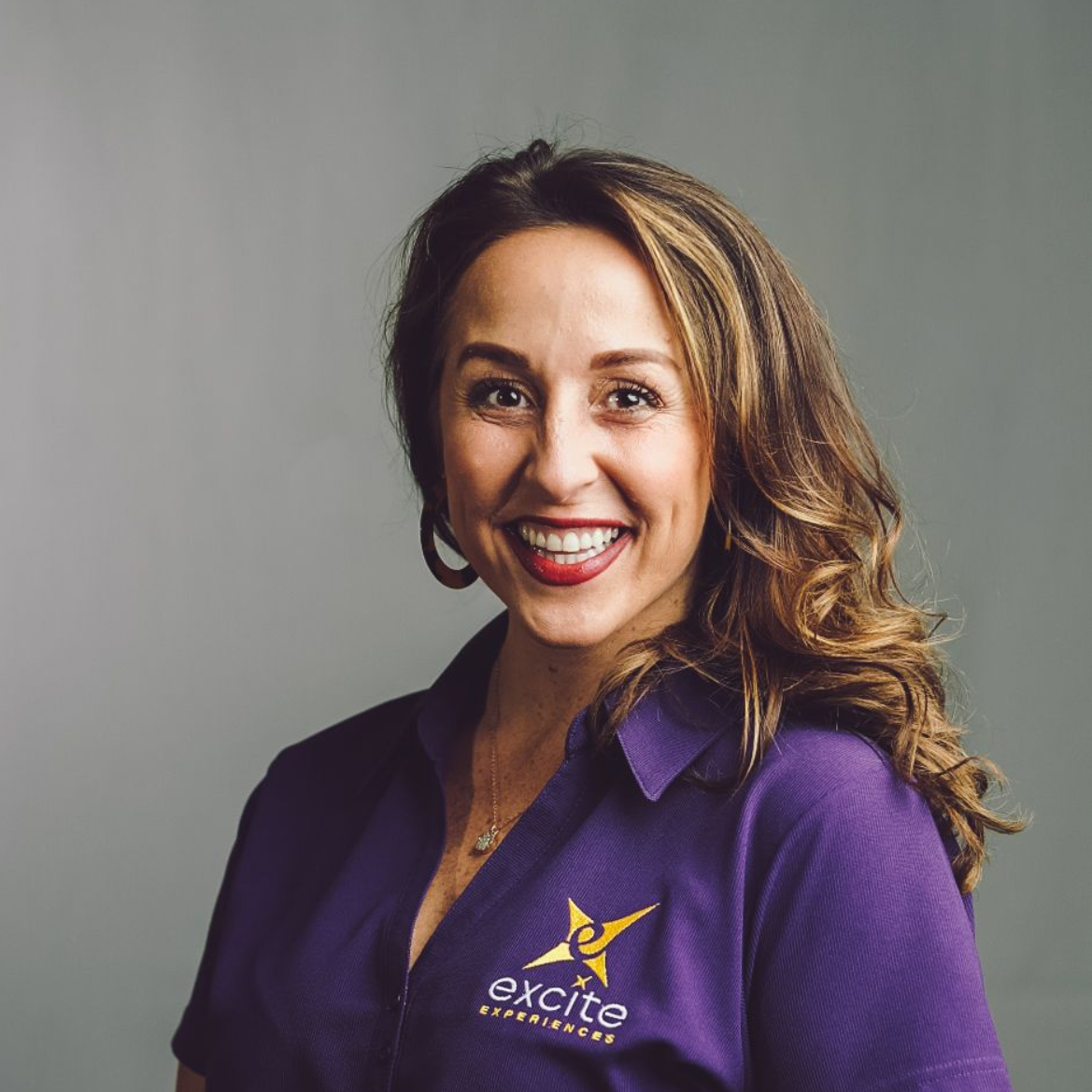 Woman with long, wavy brown hair smiles, wearing a purple shirt with a logo, against a gray backdrop.