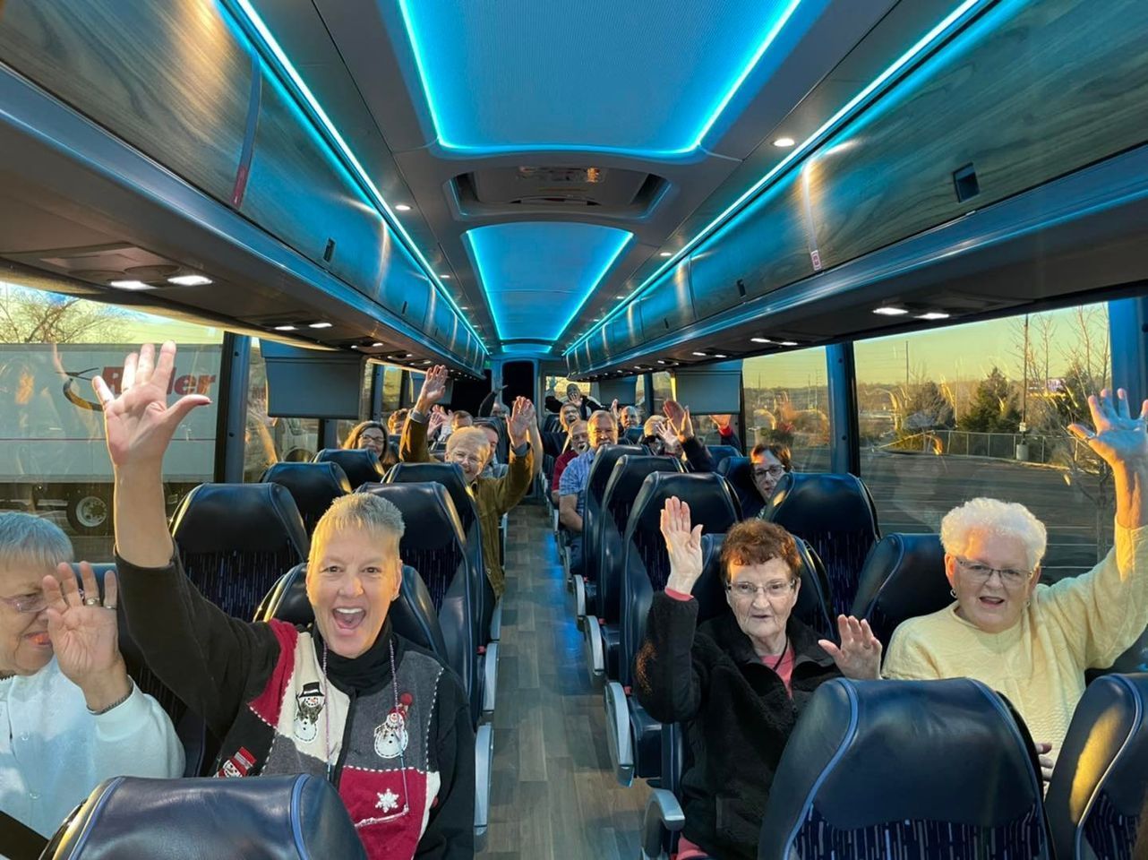 People waving from inside a bus with blue interior lighting. Sunlight streams through windows.