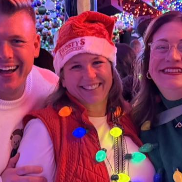Three smiling people pose in a festive setting with holiday lights. One wears a Santa hat and vest, another smiles.