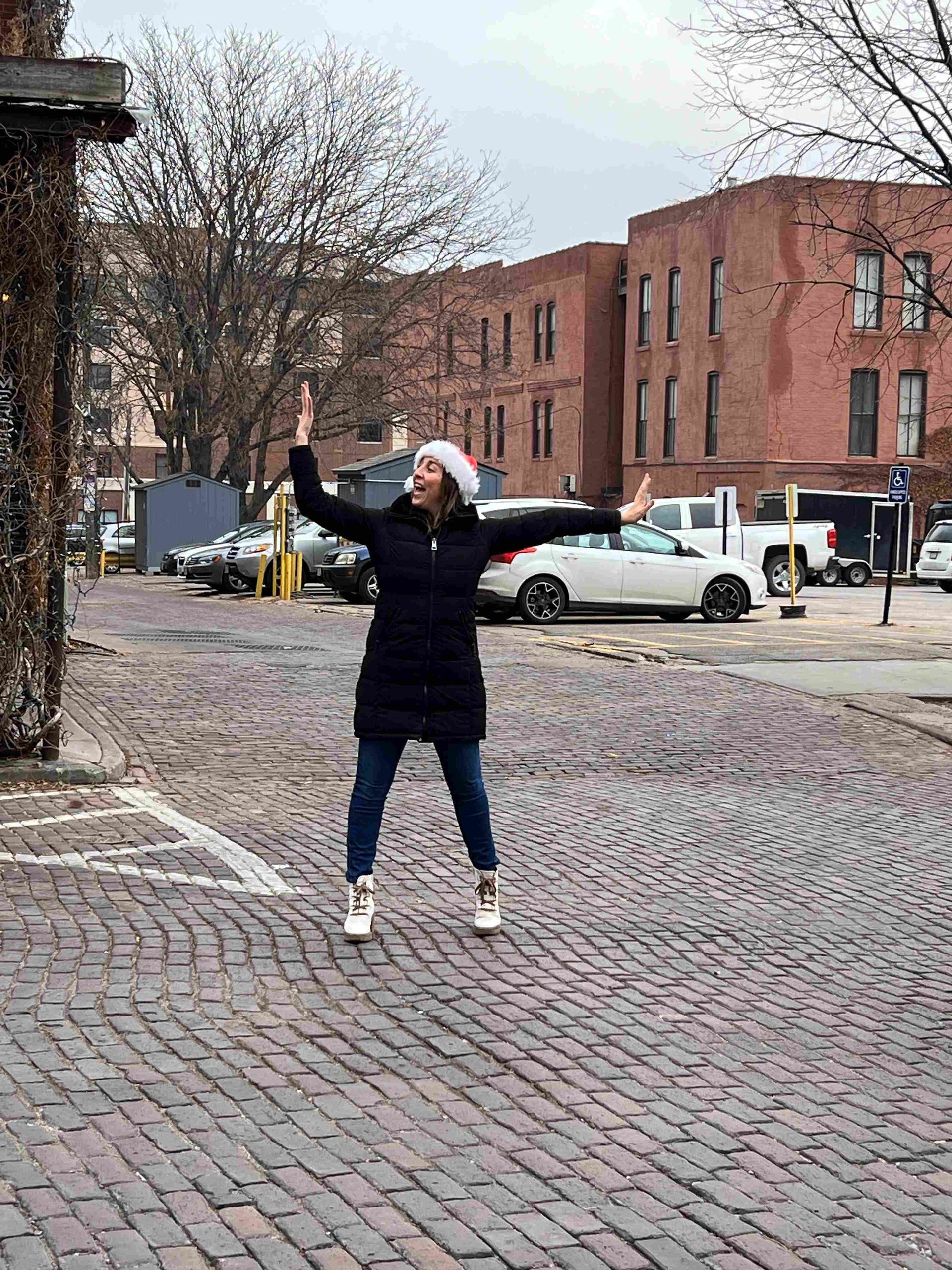 A woman is standing on a cobblestone street with her arms outstretched.