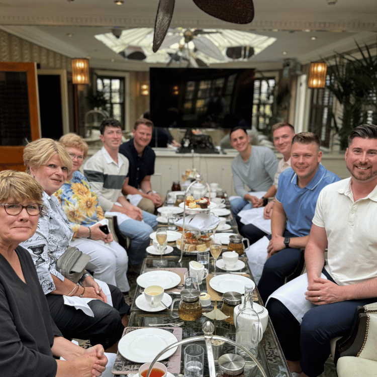 A group sits around a table set for tea in a well-lit room. They are smiling, engaged in conversation.