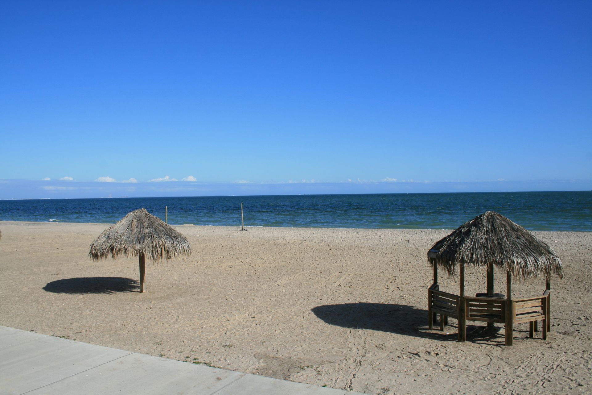 Sandy beach with two thatched-roof structures under a clear blue sky. The ocean is visible in the background.
