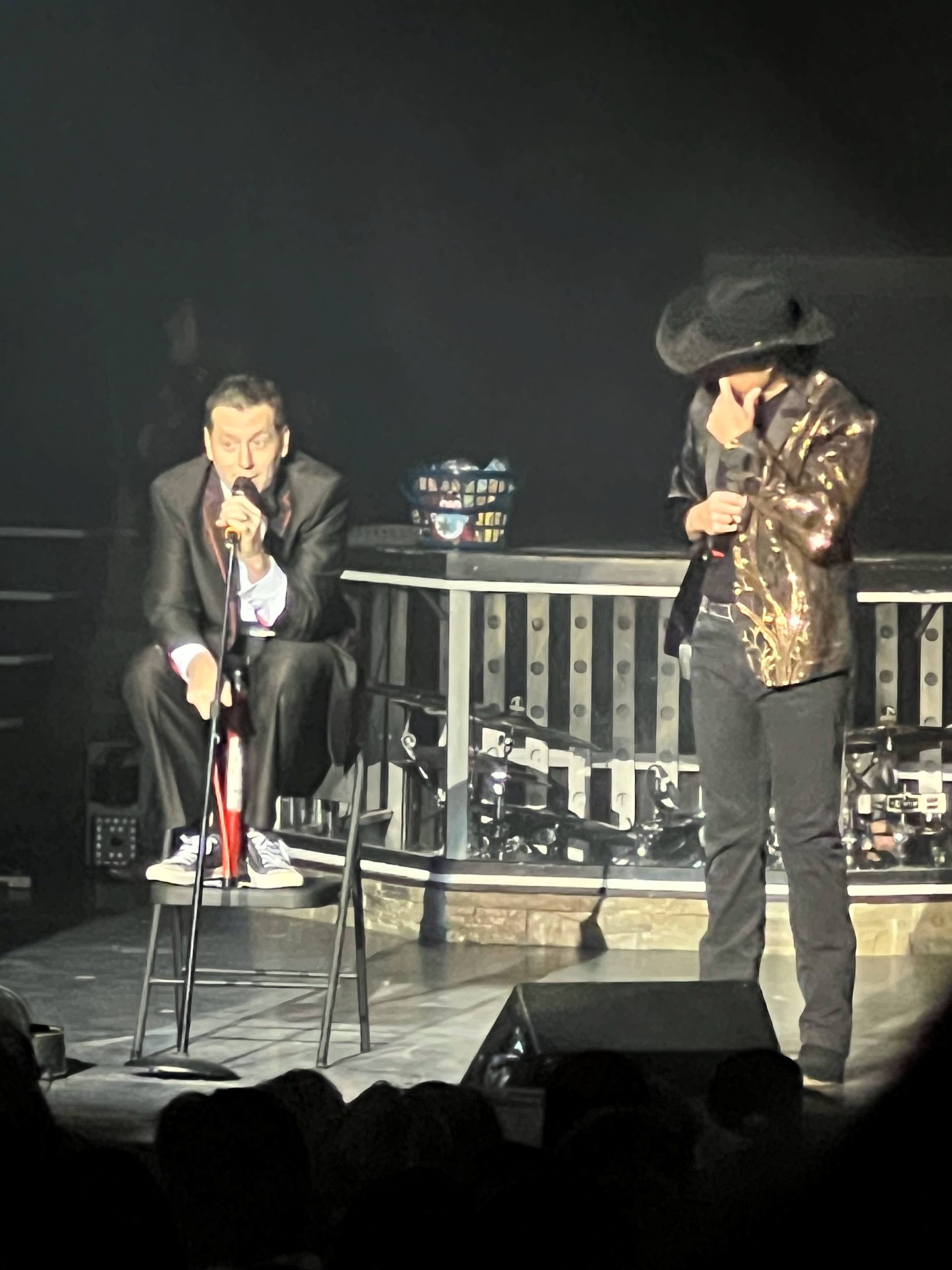 Two men singing into microphones on a stage with one wearing a cowboy hat