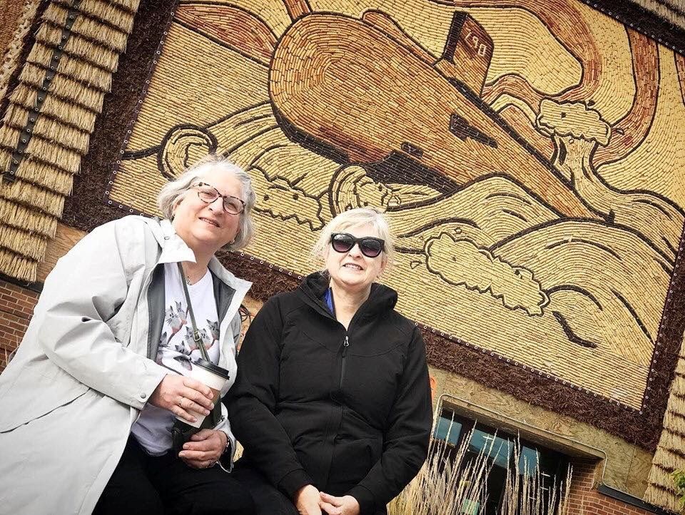 Two women are standing in front of a mosaic wall.