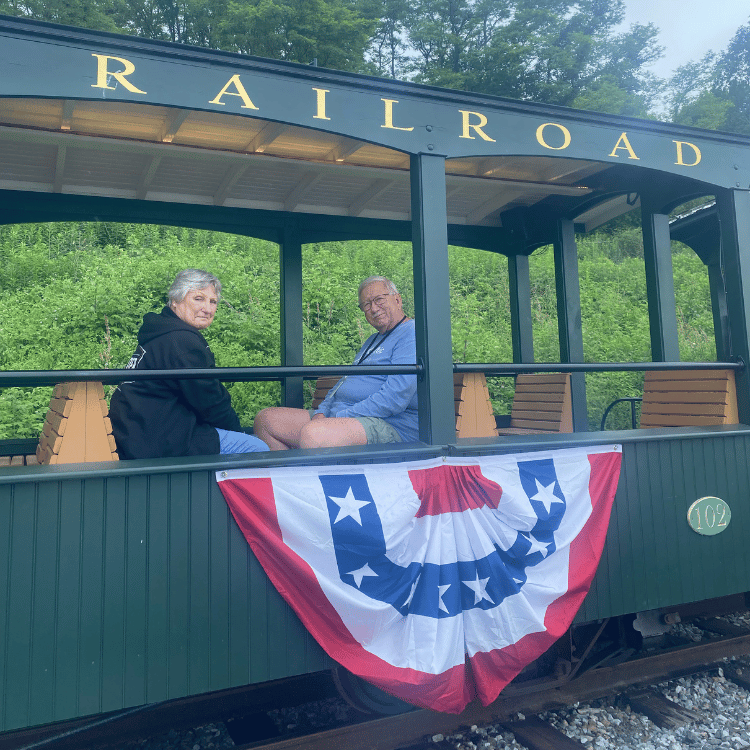 Two people sit in an open-air railroad car decorated with a patriotic banner. Green car, trees in background.