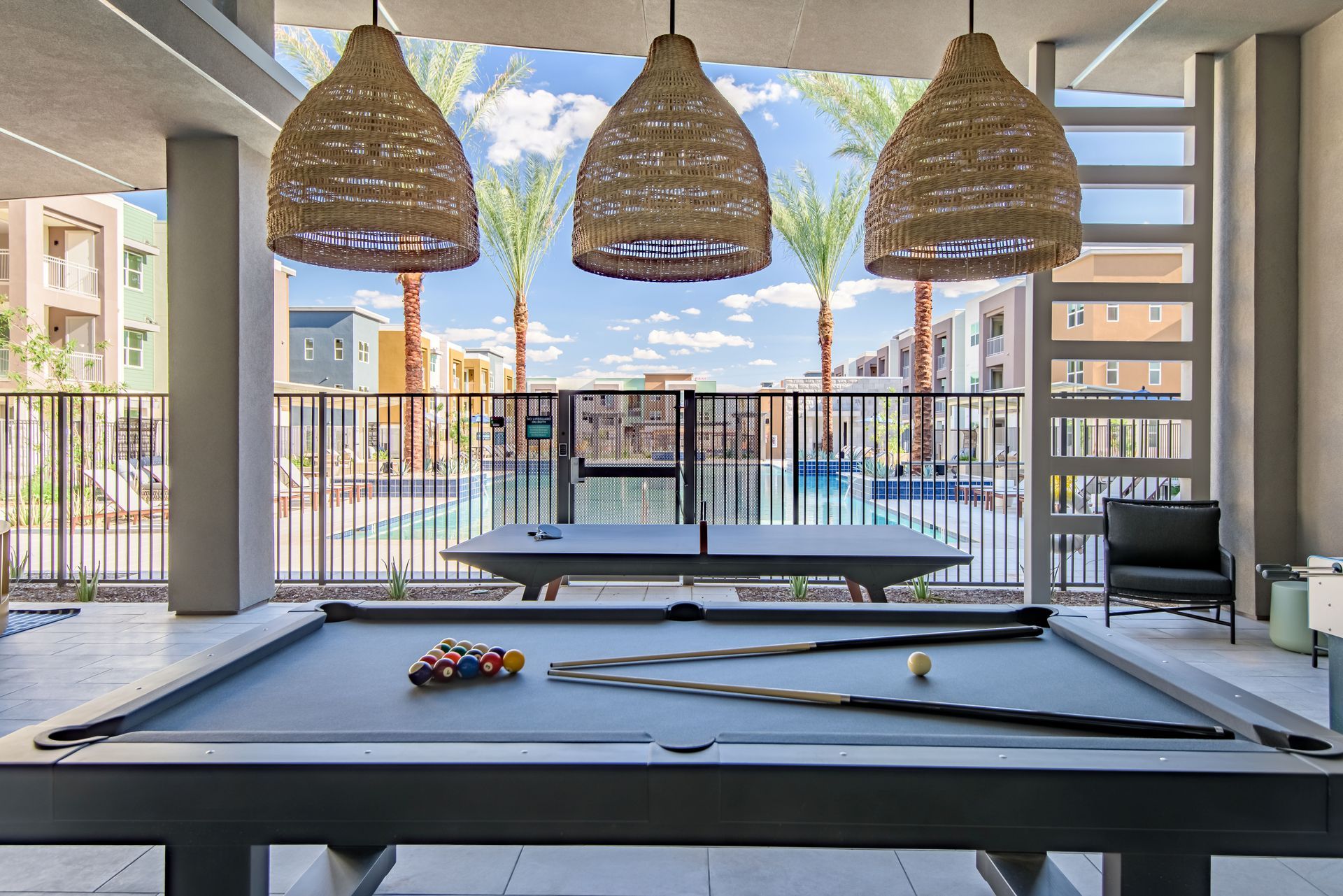 A pool table under three woven pendant lights, overlooking a swimming pool and buildings at Origin, which offers apartments for rent in Goodyear, AZ.
