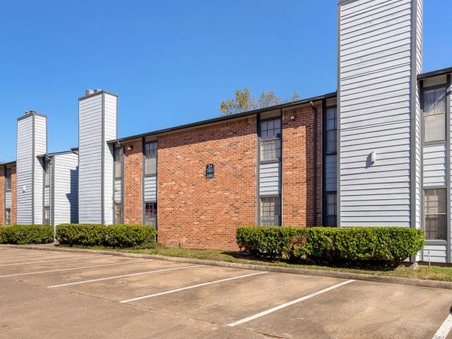 Exterior view of a brick apartment building with a parking lot and trimmed hedges.
