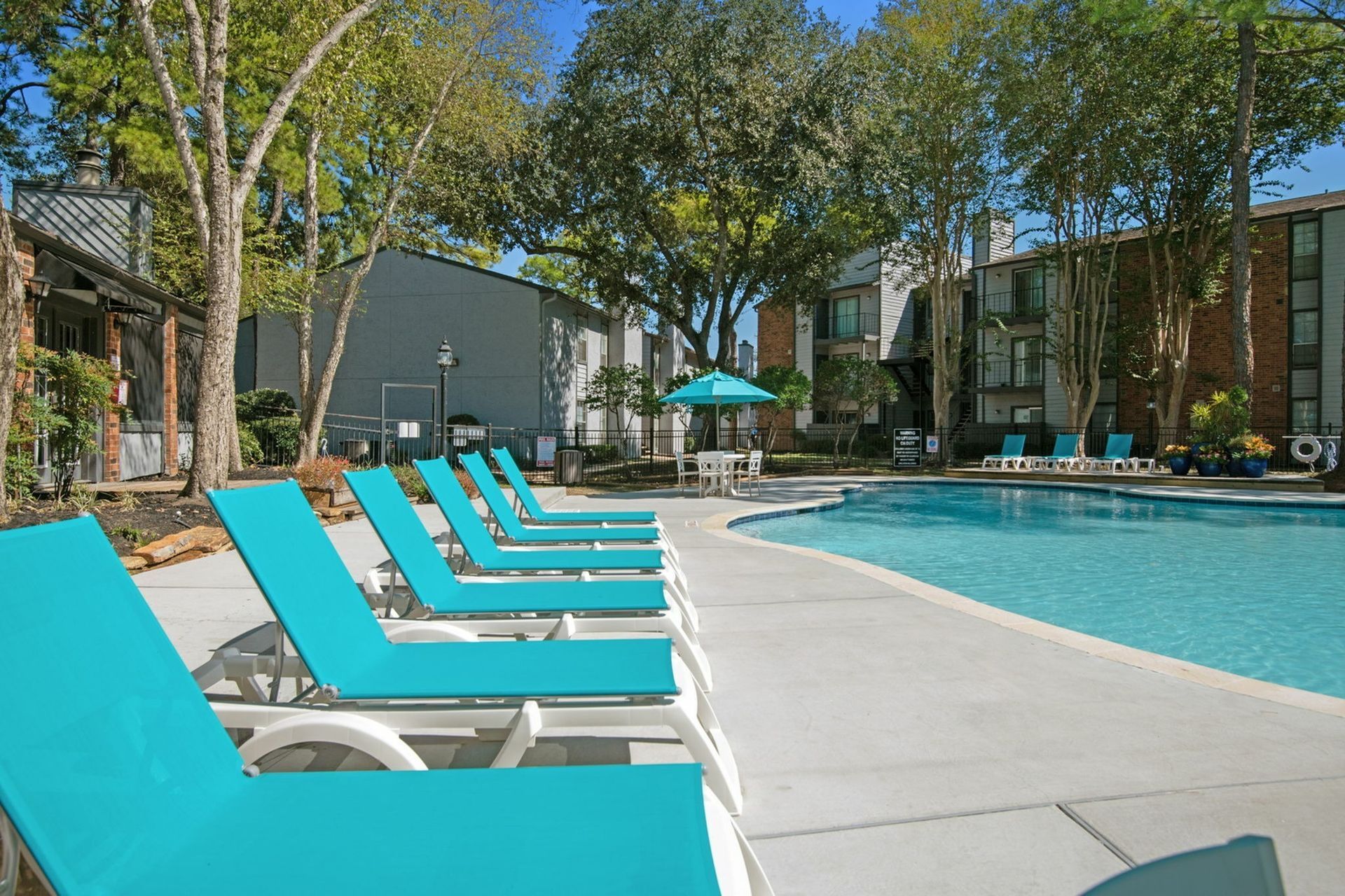 Outdoor pool area with turquoise lounge chairs, trees, and surrounding apartment buildings.
