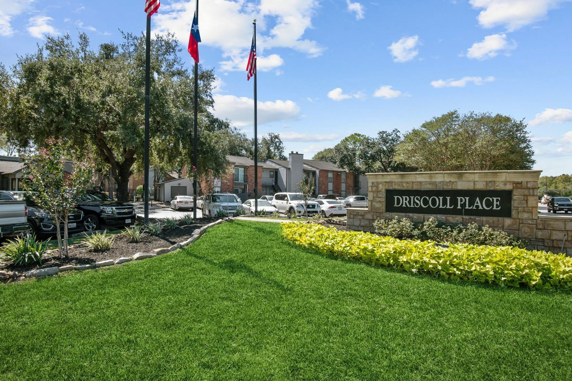 Exterior view of Driscoll Place entry with flagpoles, landscaping, and parked cars.