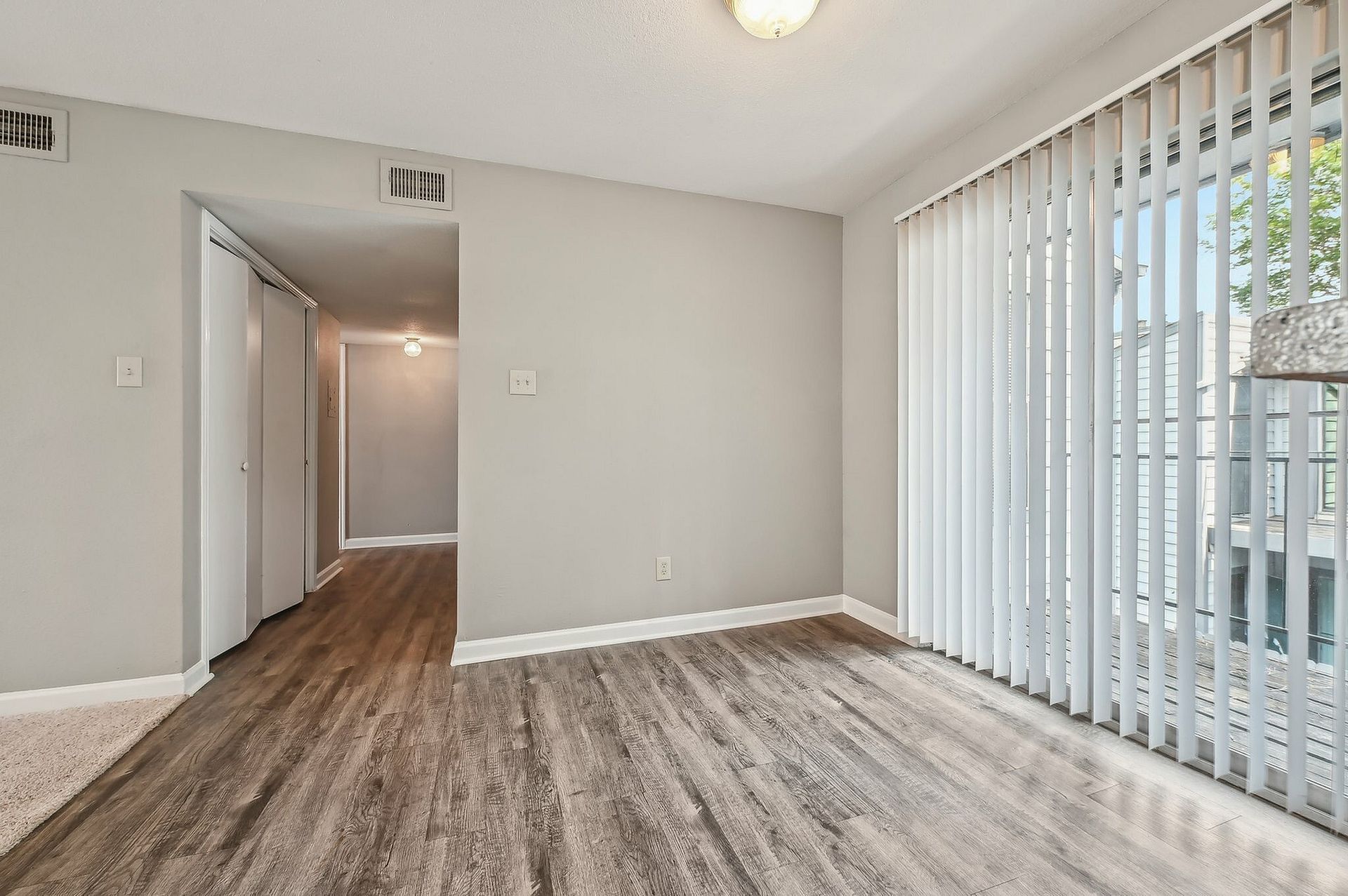 Empty apartment living area with gray walls, wood-look flooring, and a large window with vertical blinds.