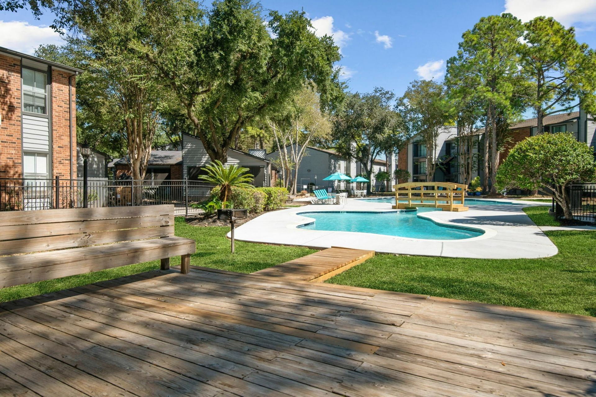 Outdoor pool area at an apartment community with lounge chairs, umbrellas, and surrounding trees.