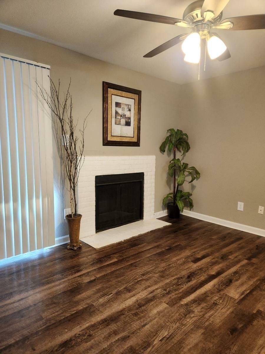 Living room with a white brick fireplace, ceiling fan, wood-look floors, vertical blinds, and a tall plant.