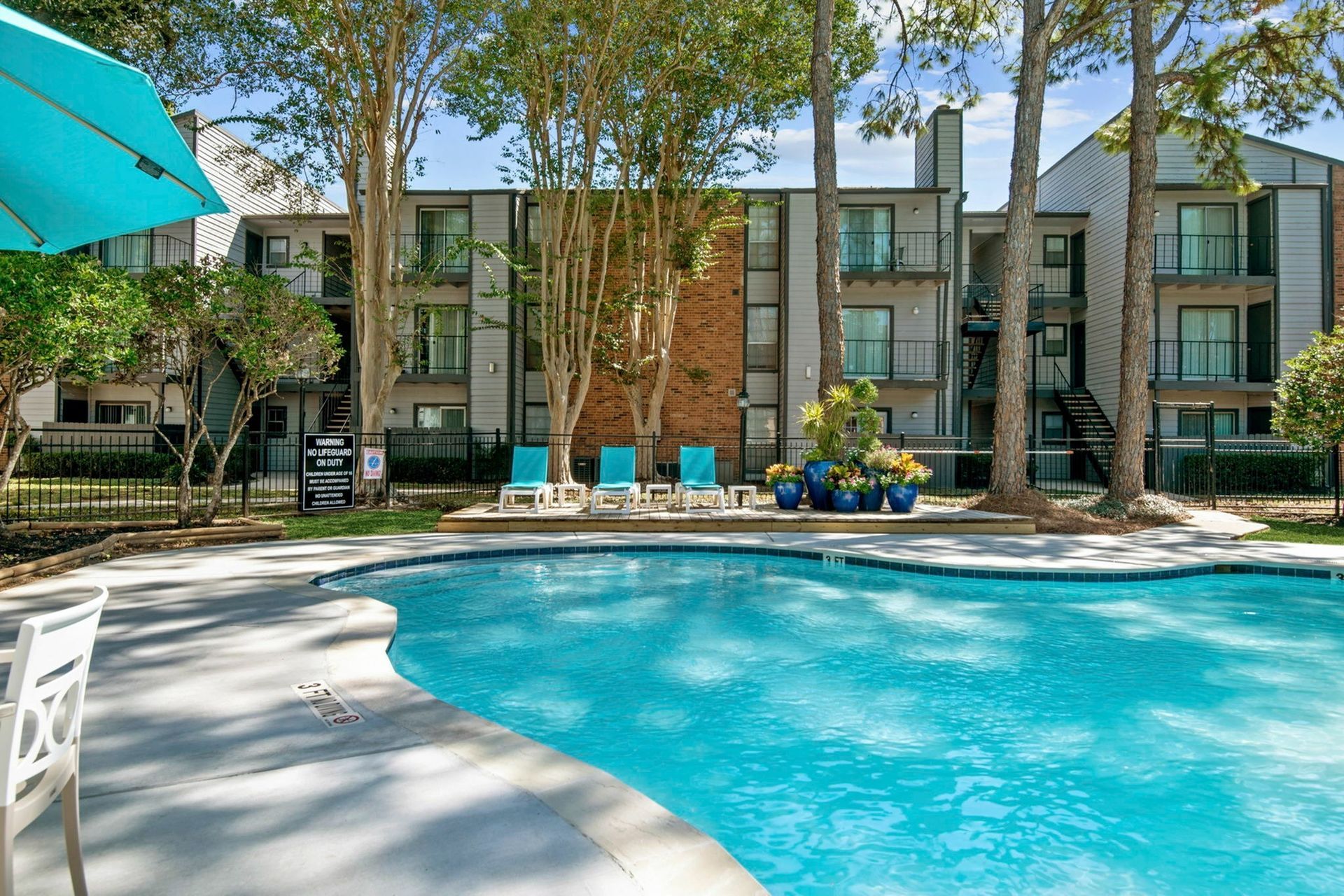 Outdoor pool with turquoise water, lounge chairs, and trees at an apartment community.
