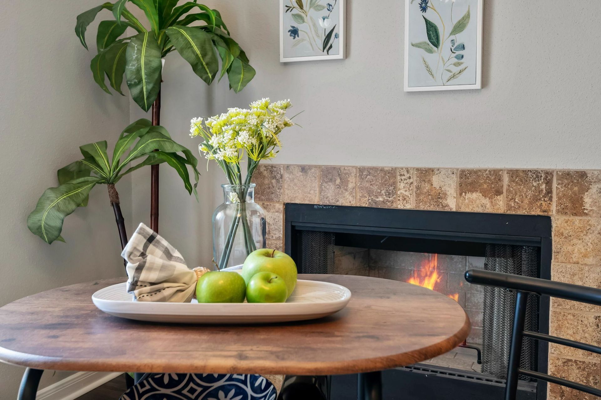 Round wooden table with a vase of white flowers and green apples beside a fireplace.