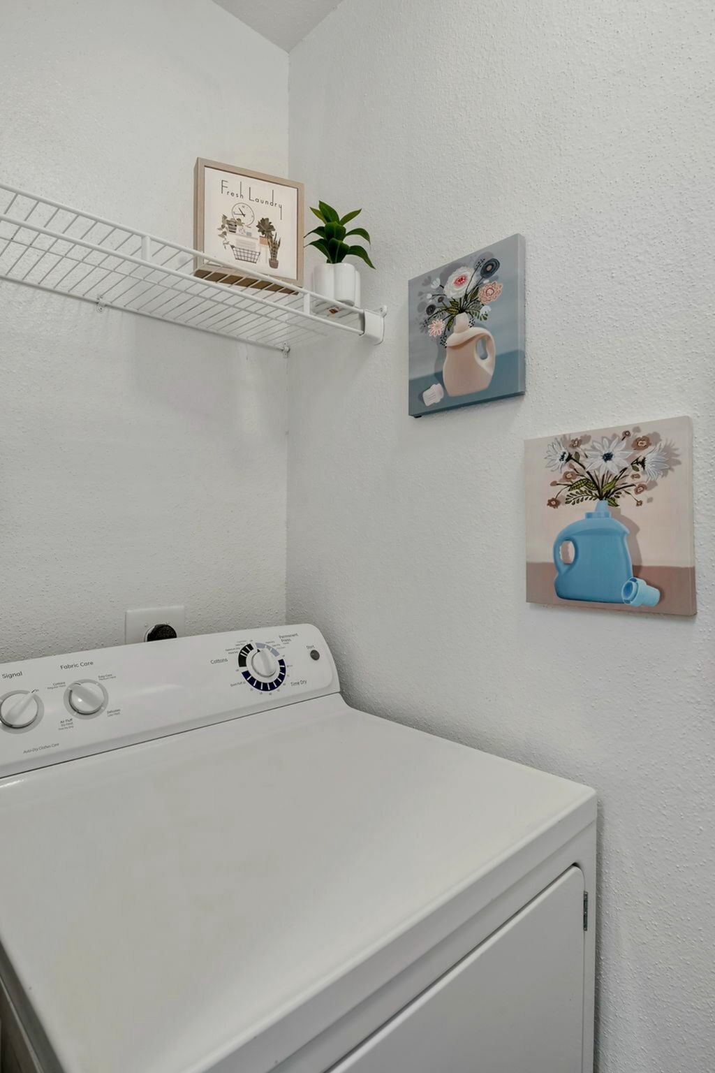 Laundry room with a top-loading washing machine, a wire shelf, and wall art.