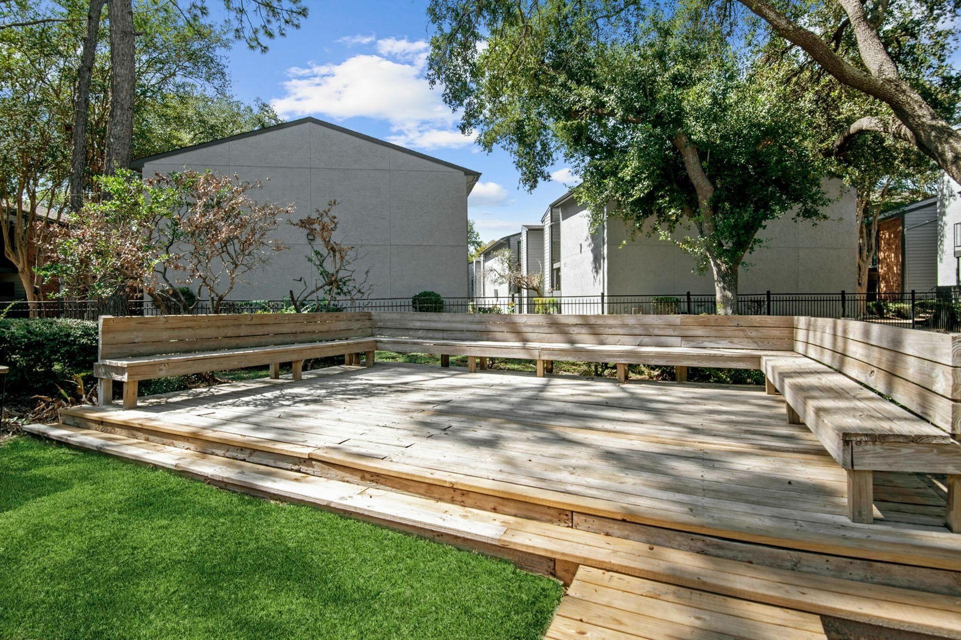 Wooden U-shaped deck seating in a courtyard with trees and apartment buildings in the background.