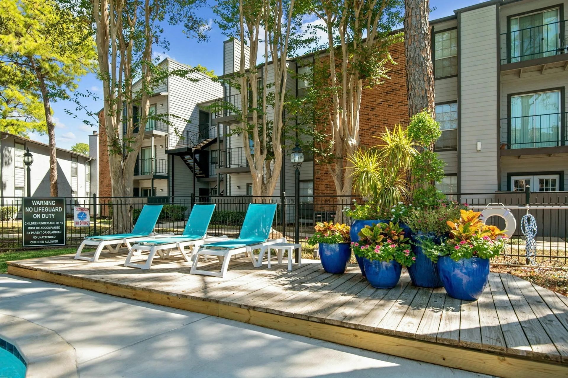 Poolside deck with turquoise lounge chairs and blue flower pots in front of a building at Driscoll Place in Houston, TX.