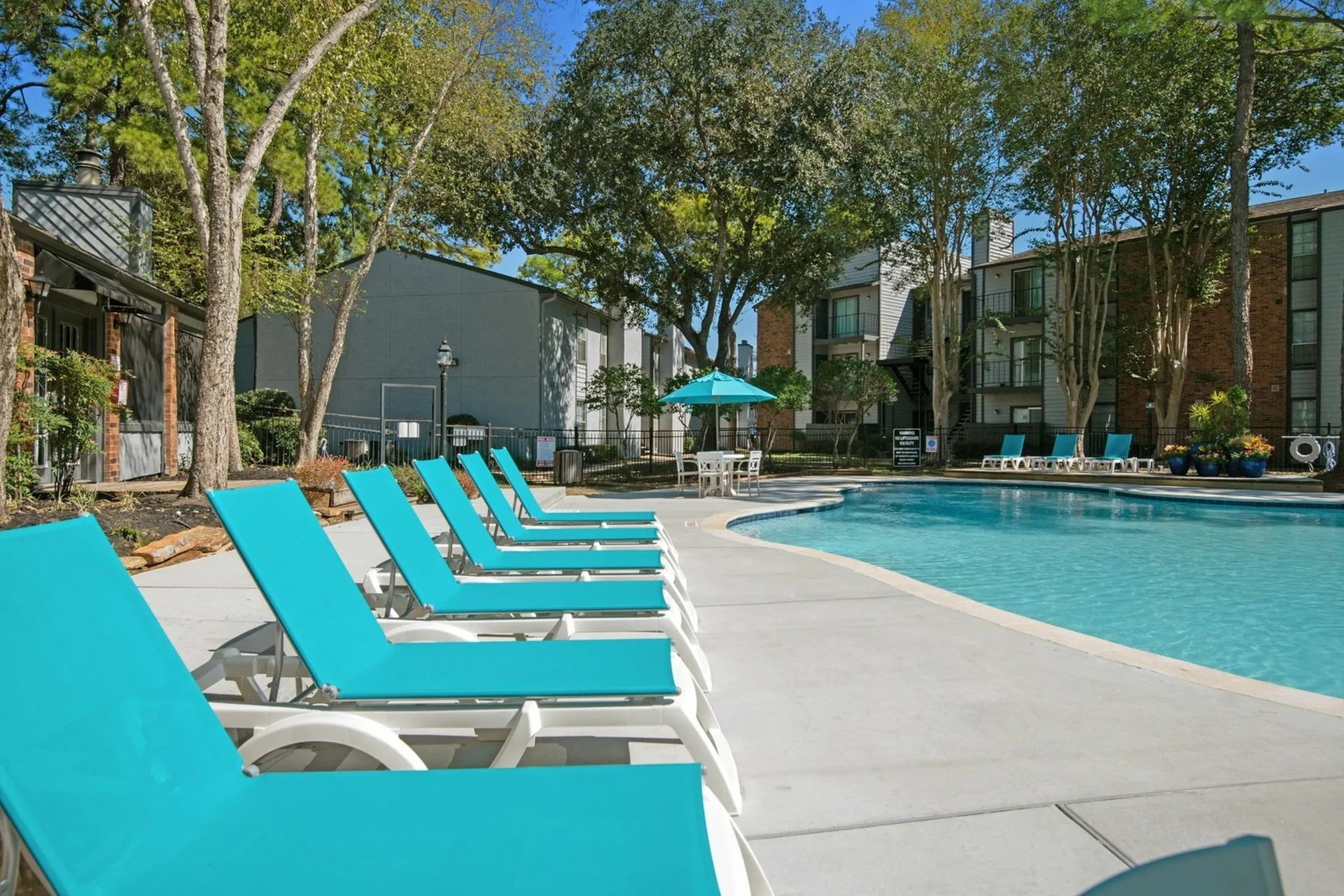 Lounge chairs by a swimming pool at an apartment complex. Turquoise chairs and umbrella, sunny day at Driscoll Place in Houston, TX.