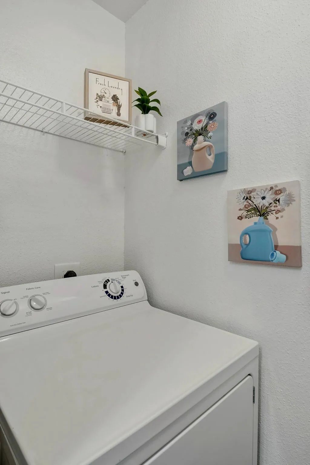 White laundry room with a dryer, a shelf with decor, and floral paintings at Driscoll Place in Houston, TX.