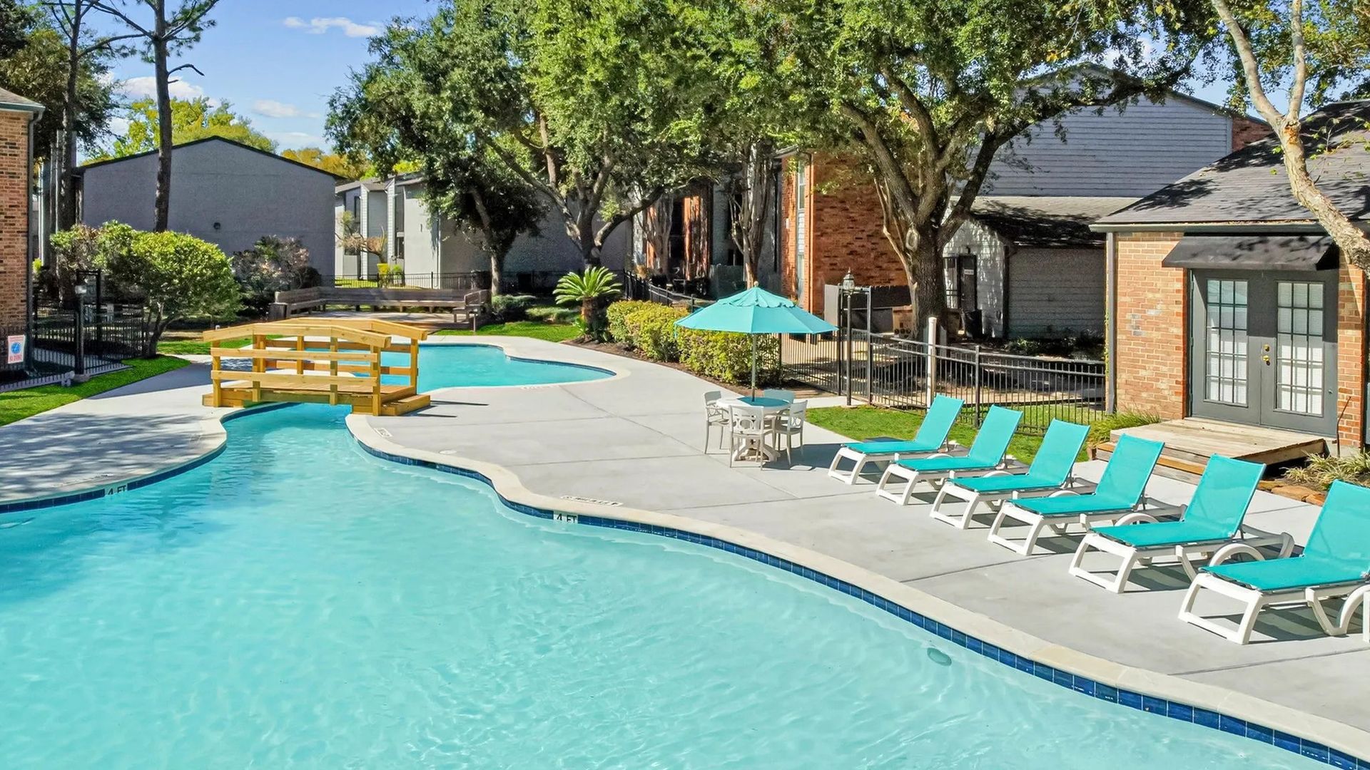 Pool with turquoise lounge chairs, umbrella, and wooden bridge, in a sunny outdoor setting at Driscoll Place in Houston, TX.