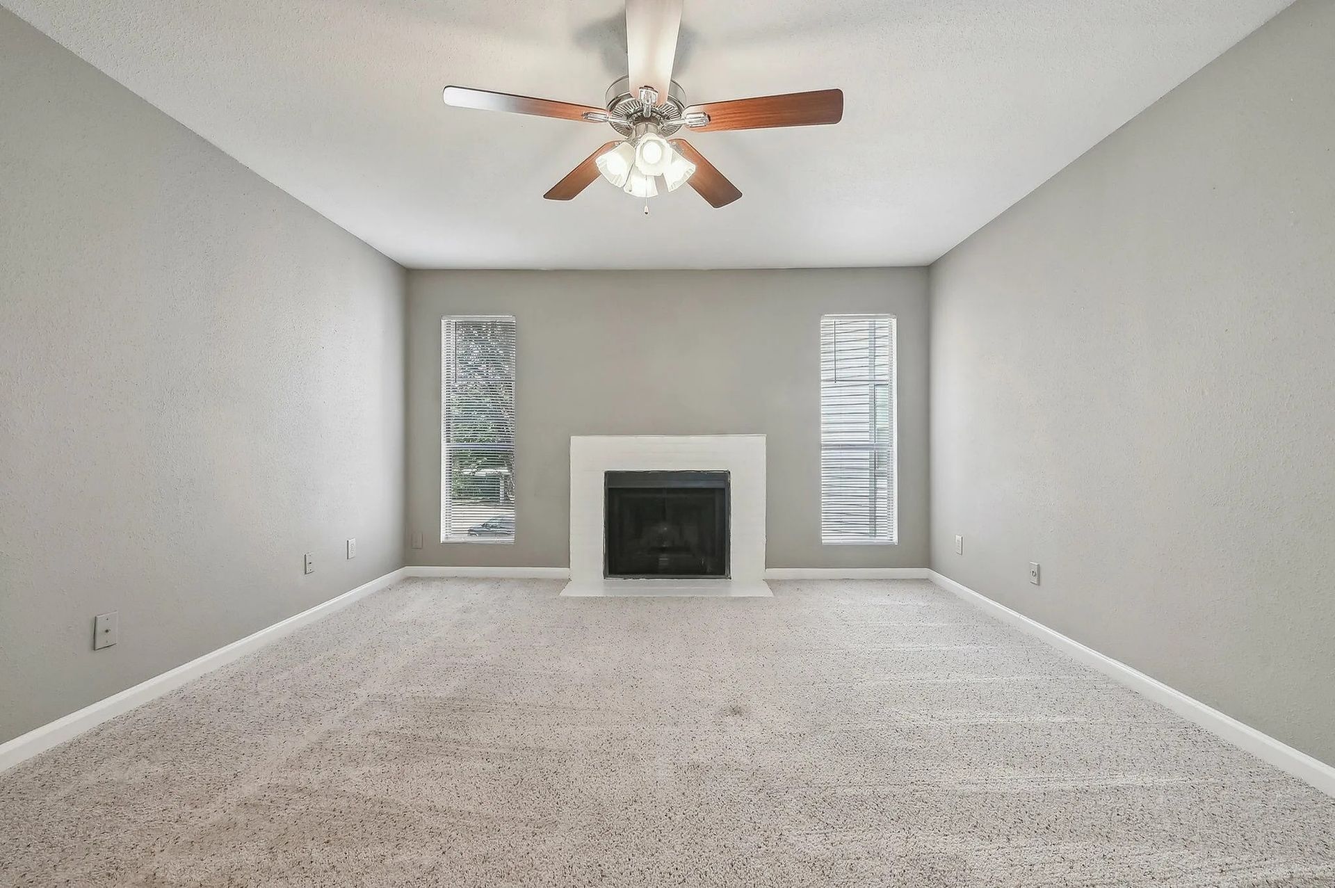 Empty living room with a fireplace, windows, a ceiling fan, and gray walls and carpet.