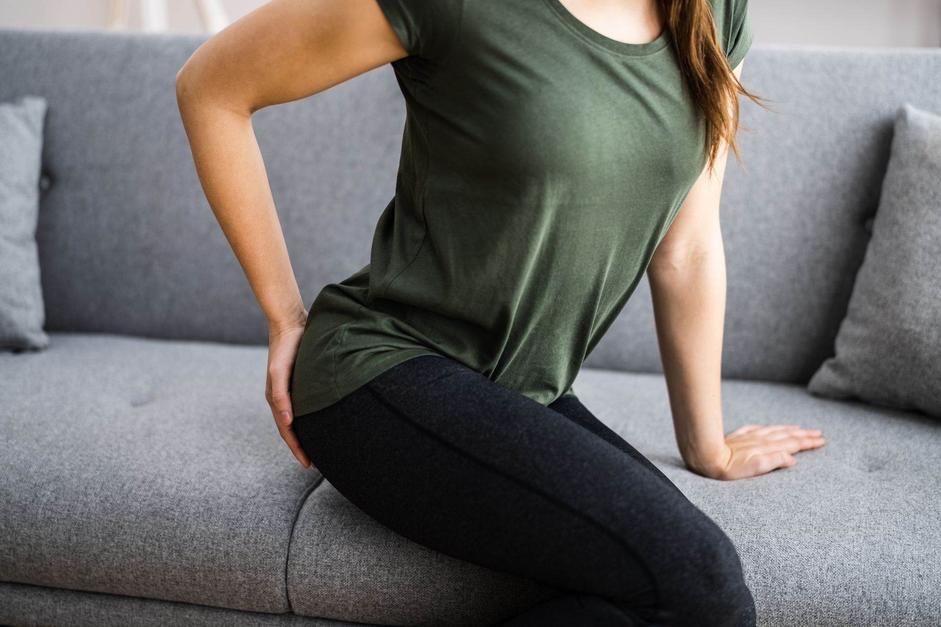 Mulher sentada no sofá, segurando a região lombar, aparentando desconforto.