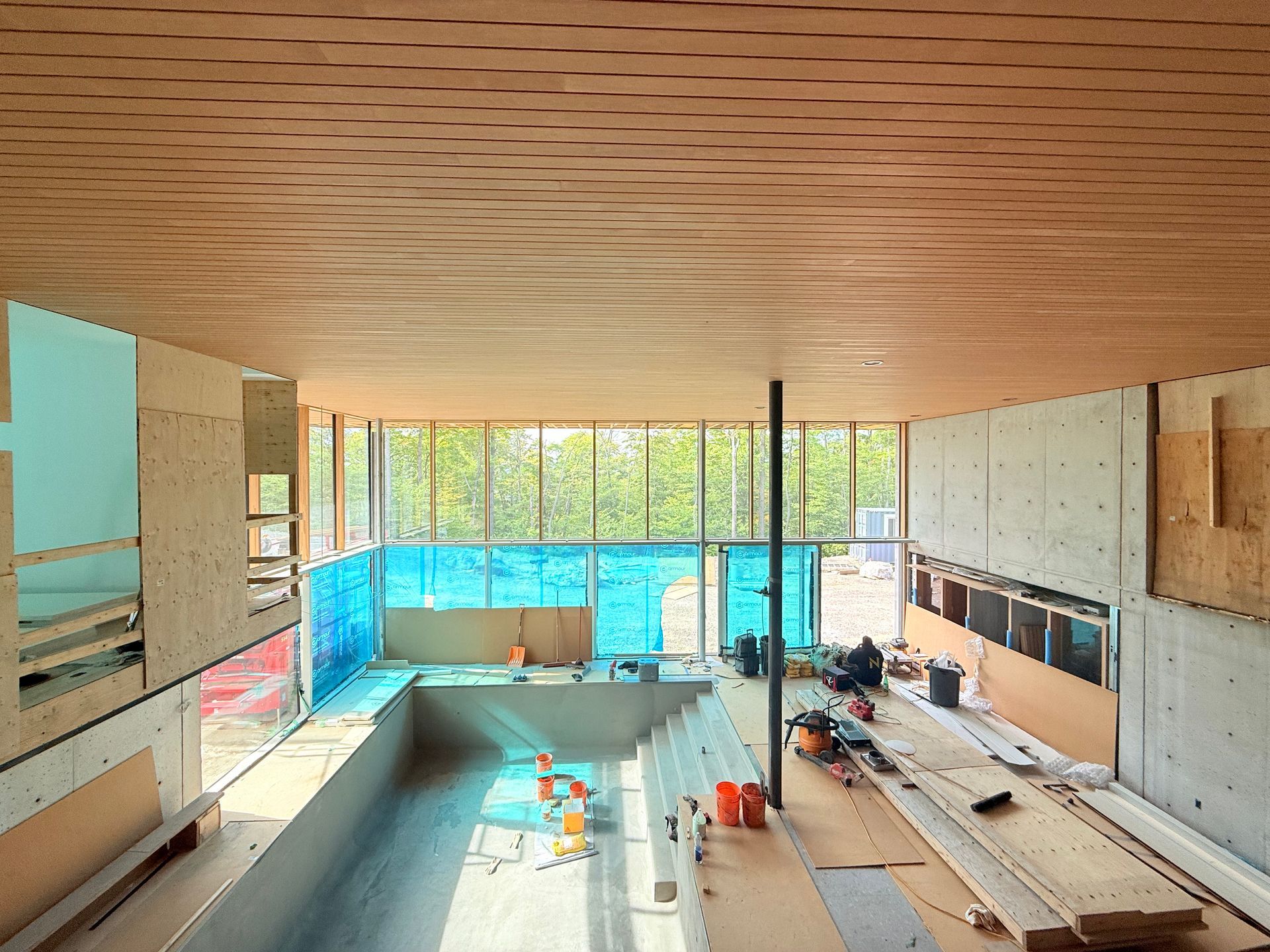 Piscine intérieure en construction ; plafond en bois, murs en béton, grandes fenêtres.