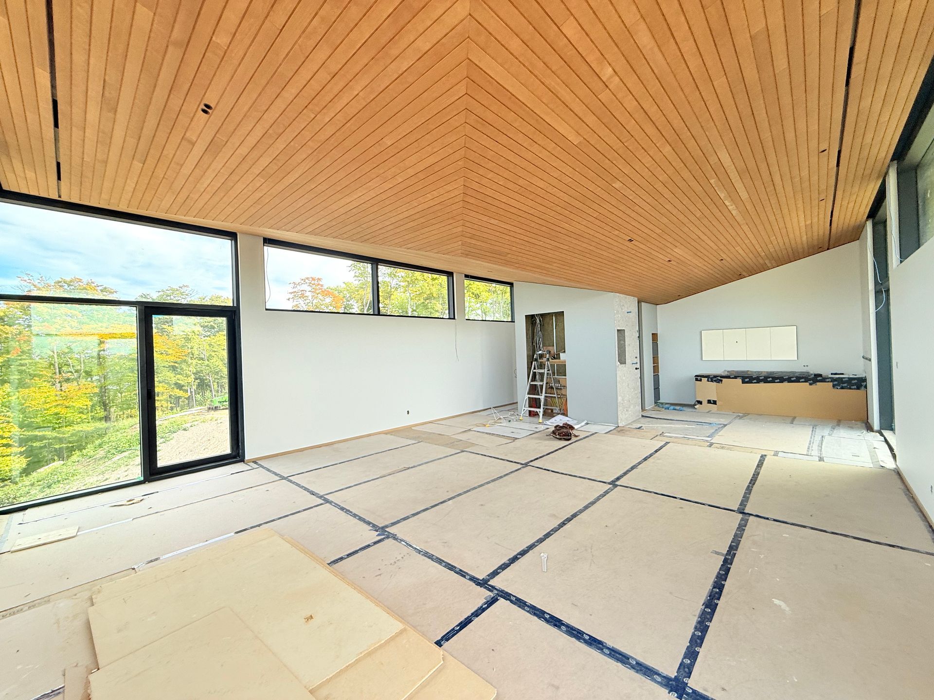 Intérieur d'une pièce en construction avec de grandes fenêtres et un plafond en bois. Le revêtement de sol est prêt à être posé.