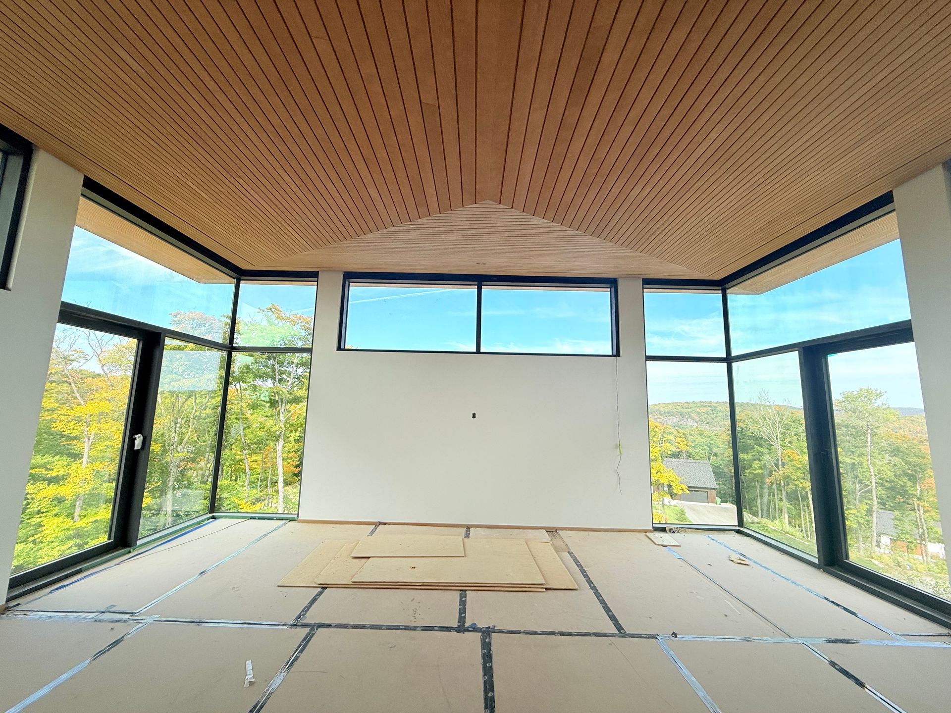Intérieur d'une pièce avec de grandes fenêtres, un plafond en bois et un sol brut. Vue sur un paysage verdoyant et un ciel bleu.