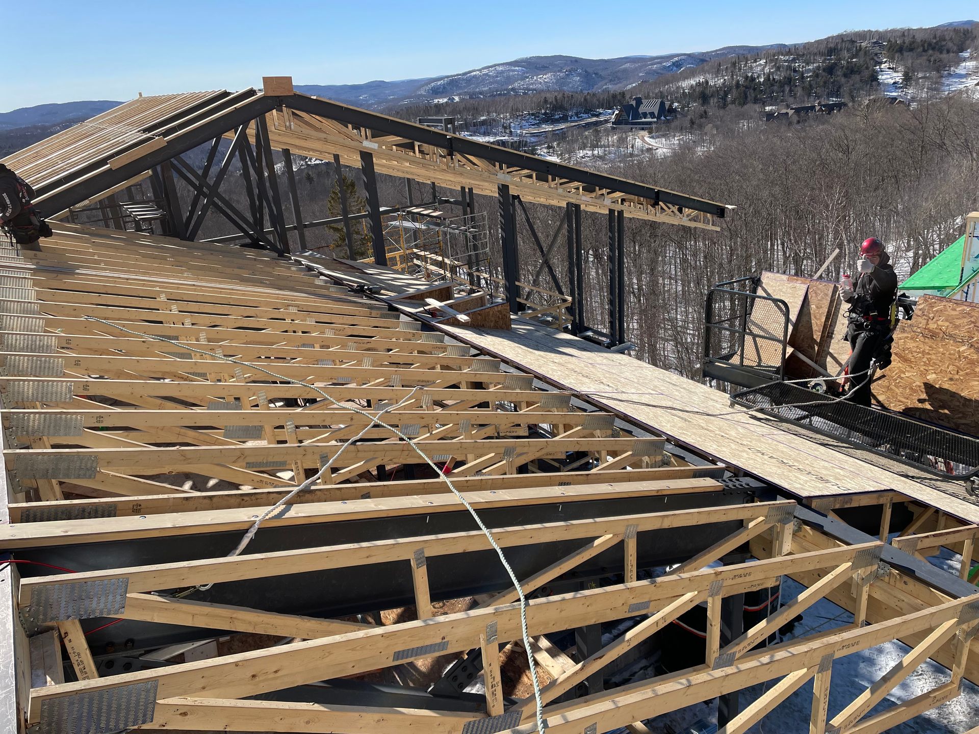 Chantier de construction : charpente de toit en bois en cours de construction, ouvrier à droite, montagne enneigée en arrière-plan.