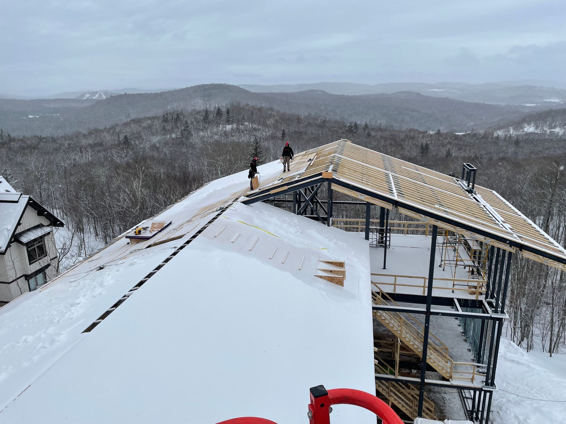 Travaux de construction sur un toit enneigé avec deux ouvriers. Charpente en bois et montagne enneigée en arrière-plan.