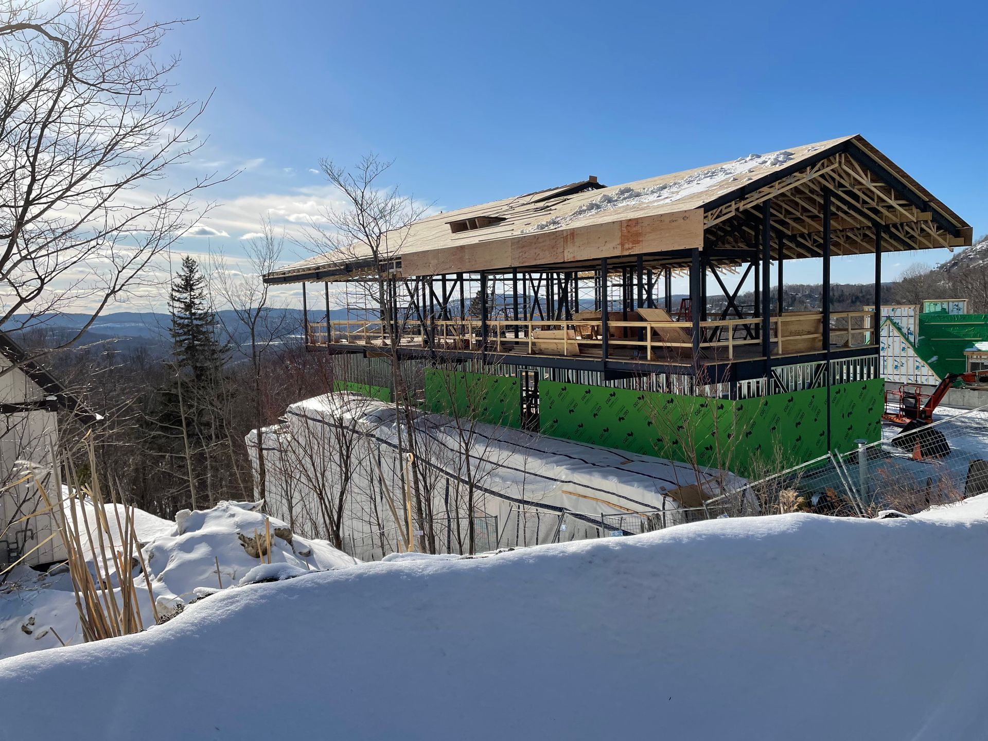 Bâtiment en construction à flanc de colline enneigée, toit inachevé et bardage vert. Ciel bleu dégagé.