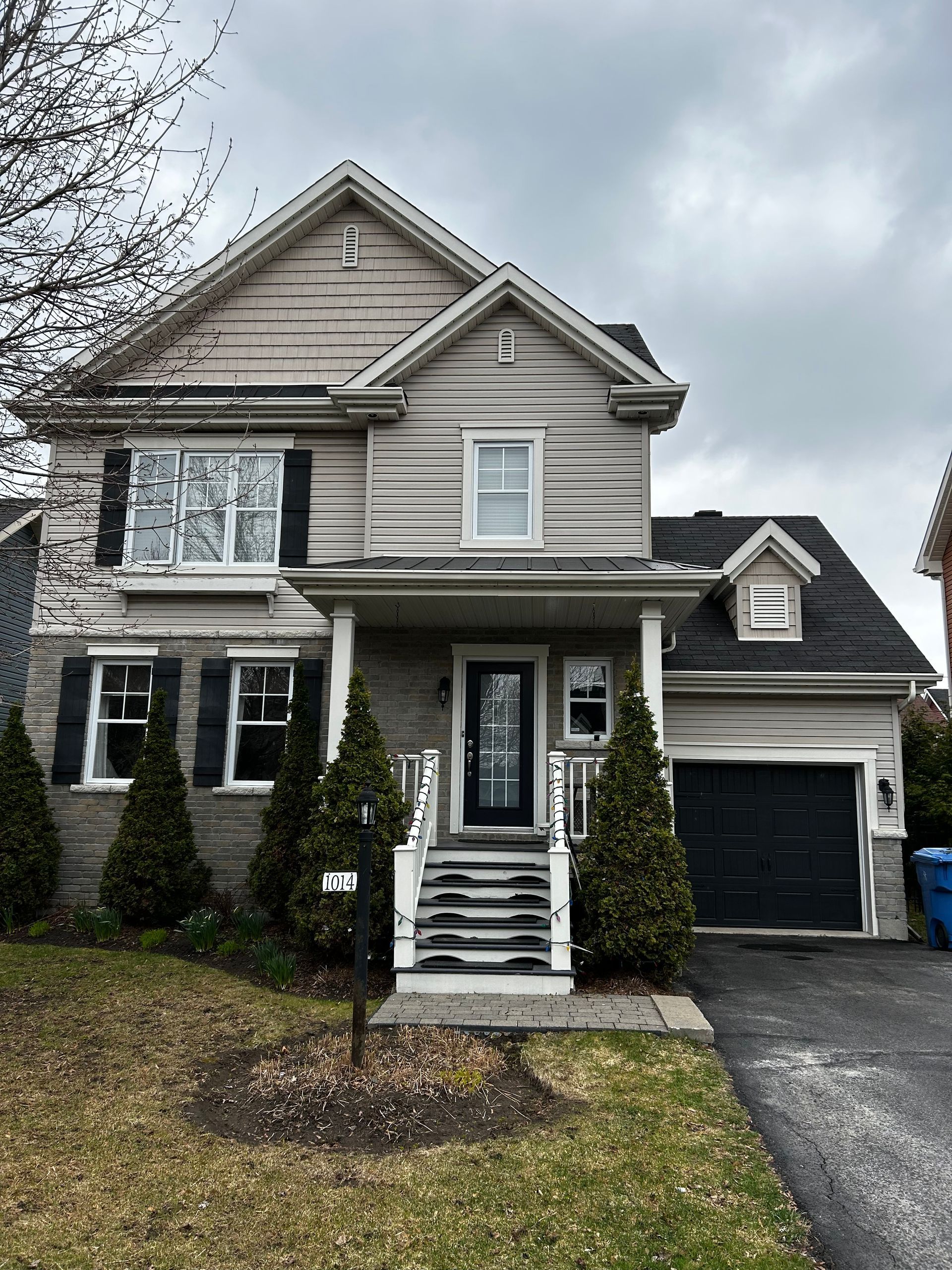 Maison à deux étages avec bardage clair, volets noirs, garage et porche sous un ciel nuageux.