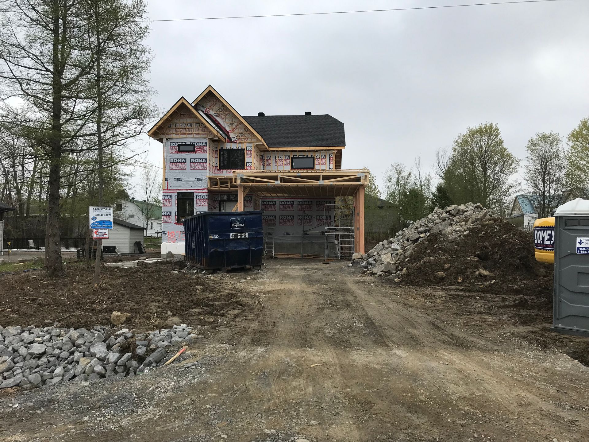 Chantier de construction avec une maison en construction, une benne à ordures et des toilettes portables.