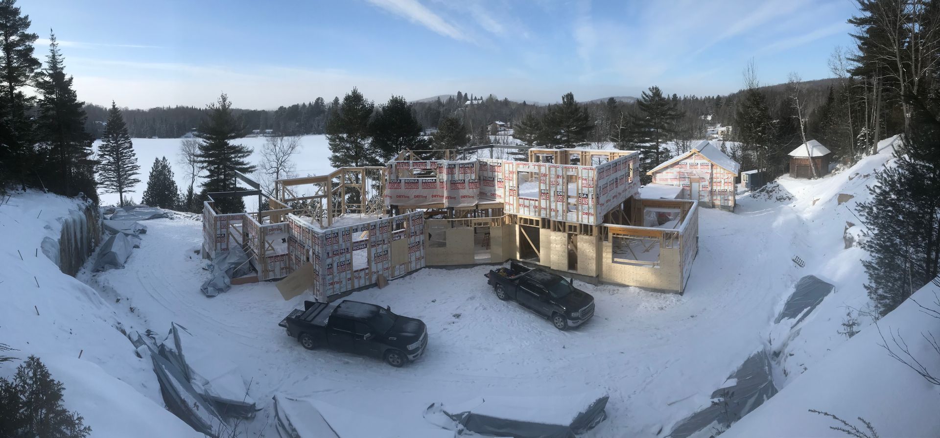 Chantier de construction entouré de neige. Bâtiments en construction, avec des arbres et un lac en arrière-plan. Deux véhicules sont présents.