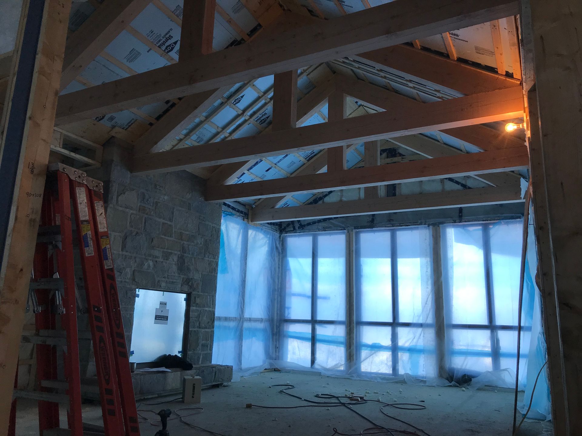 Aménagement intérieur d'une pièce, avec poutres en bois apparentes, mur en pierre, grandes fenêtres recouvertes de plastique bleu et une échelle rouge.