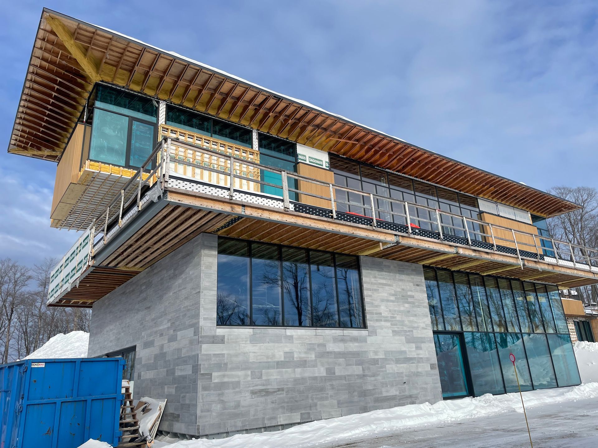 Bâtiment en construction, à deux étages, avec une base en parpaings, un toit en bois et de grandes fenêtres ; neige au sol.