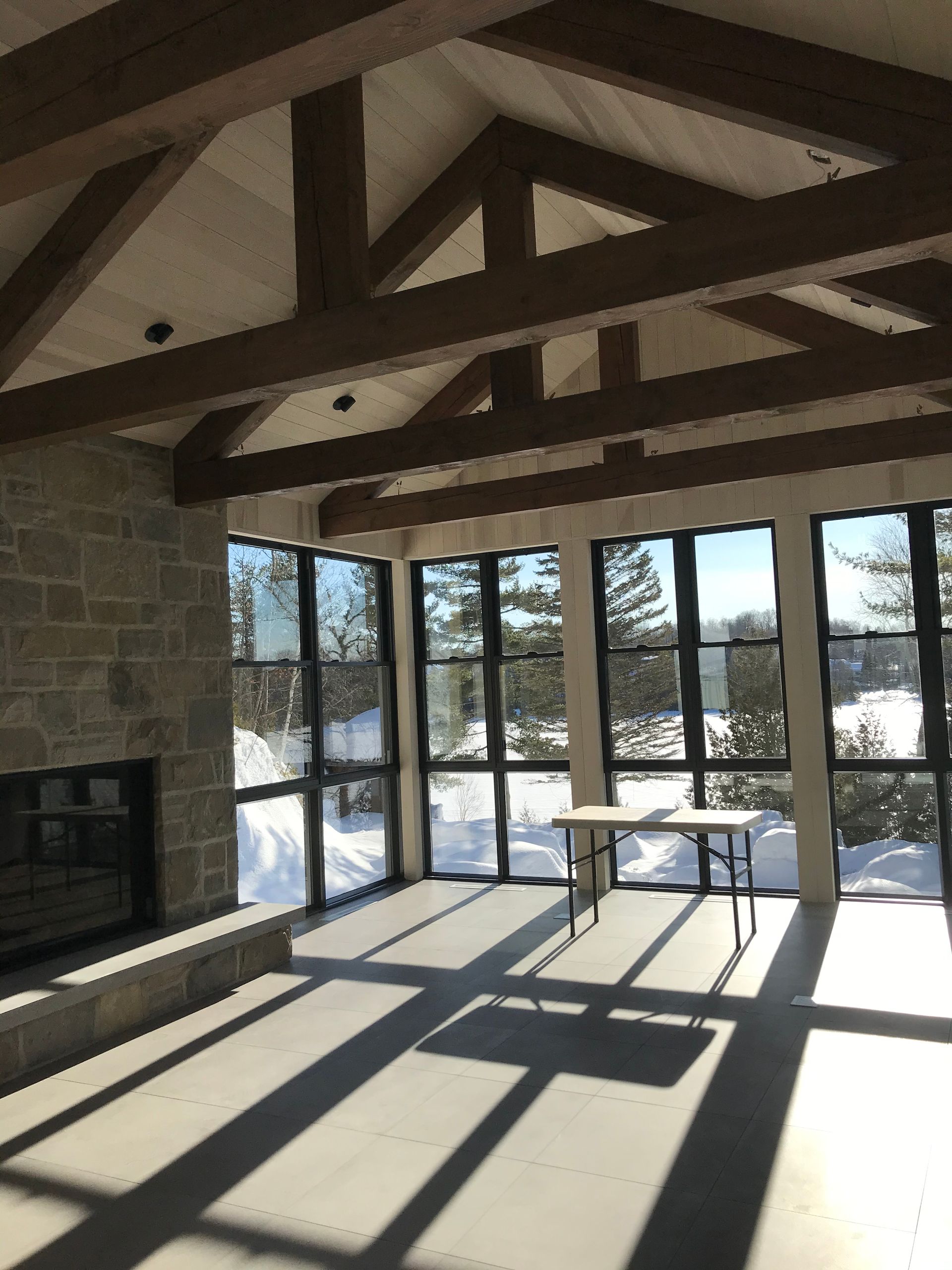 Intérieur d'une pièce avec de grandes fenêtres donnant sur un paysage enneigé. Poutres apparentes et cheminée en pierre.