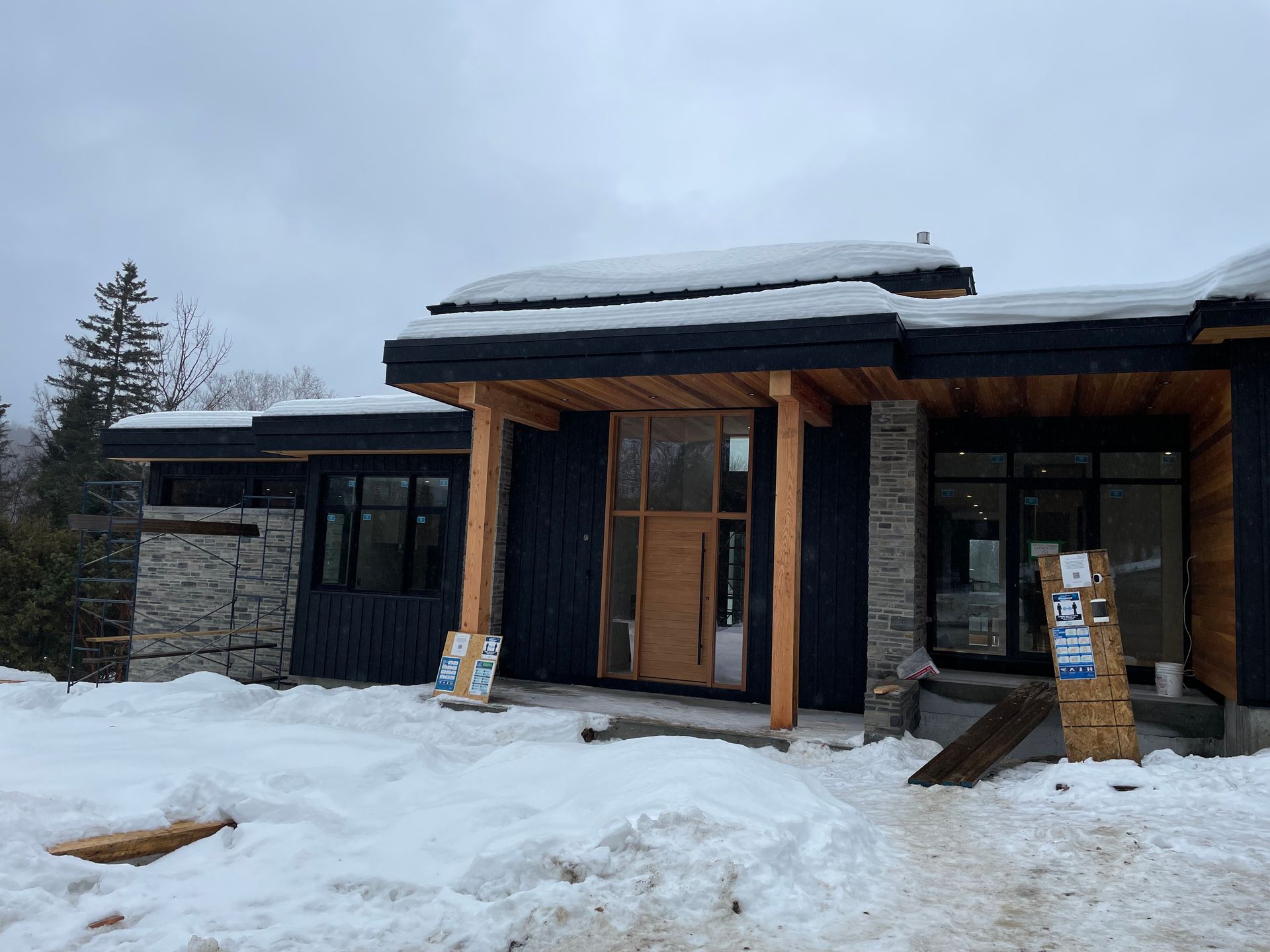 Une maison en construction, avec un bardage noir, des éléments en pierre et une entrée en bois sous un toit enneigé.