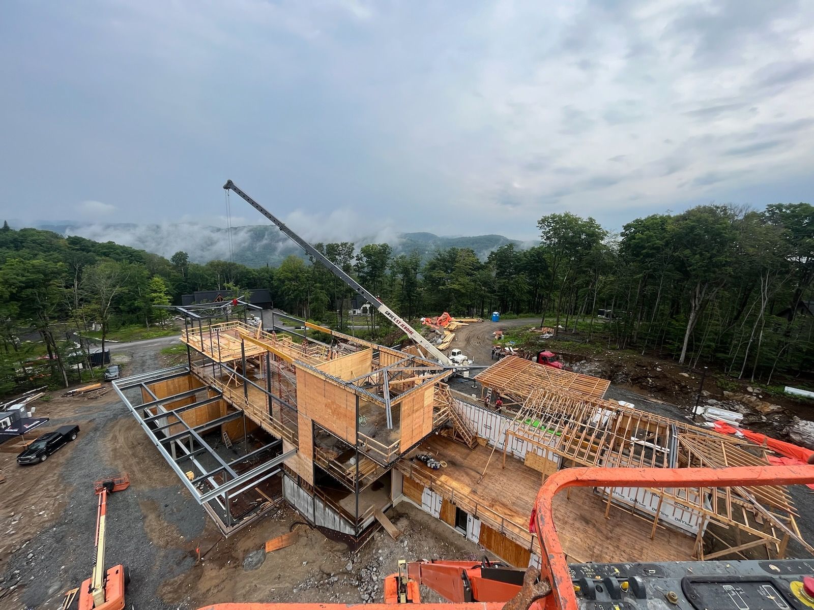 Chantier de construction avec grue, maison en bois partiellement construite, donnant sur un paysage vallonné et verdoyant sous un ciel nuageux.