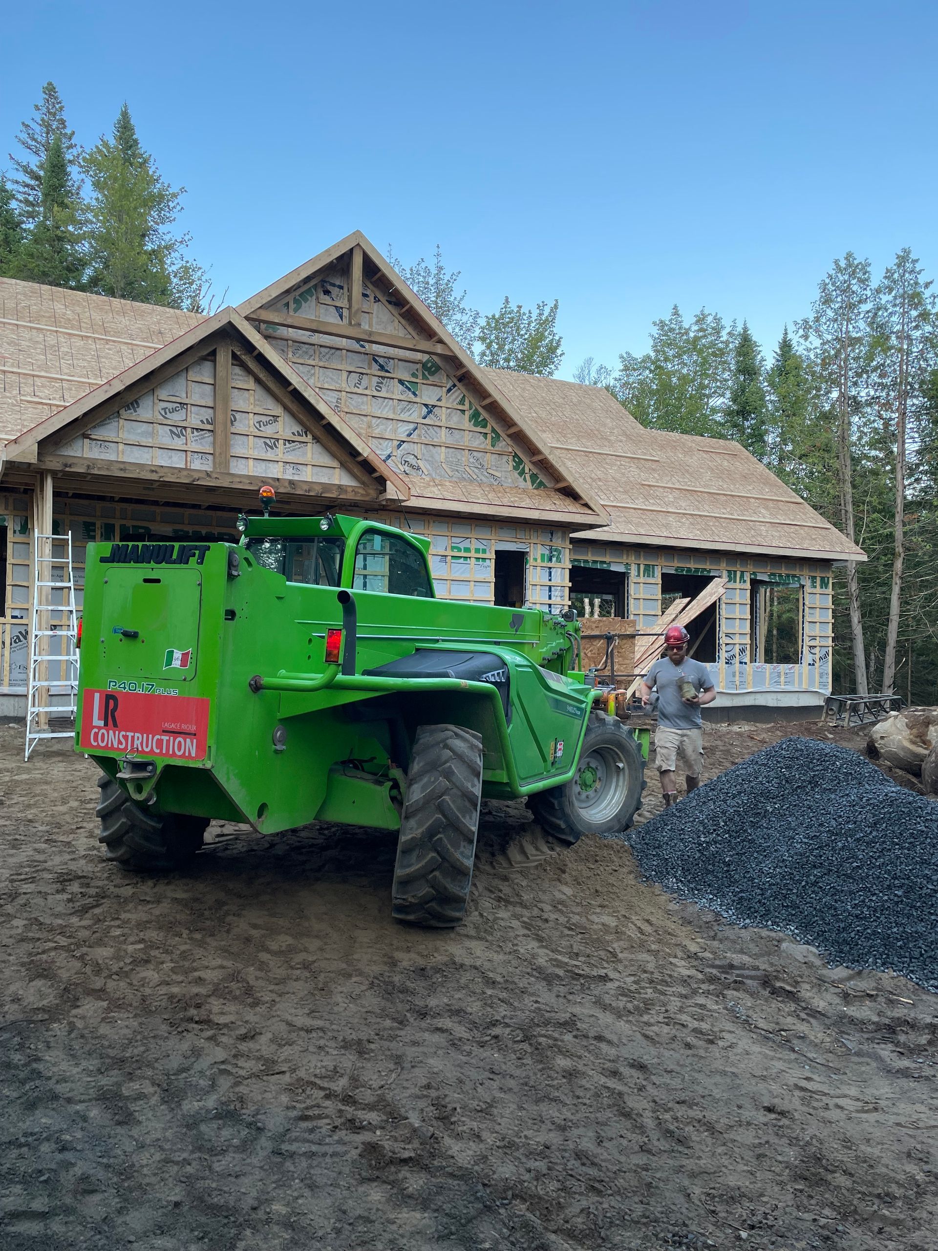 Un chariot élévateur de chantier vert se trouve près d'une maison en construction ; un ouvrier se trouve à proximité avec un tas de gravier.