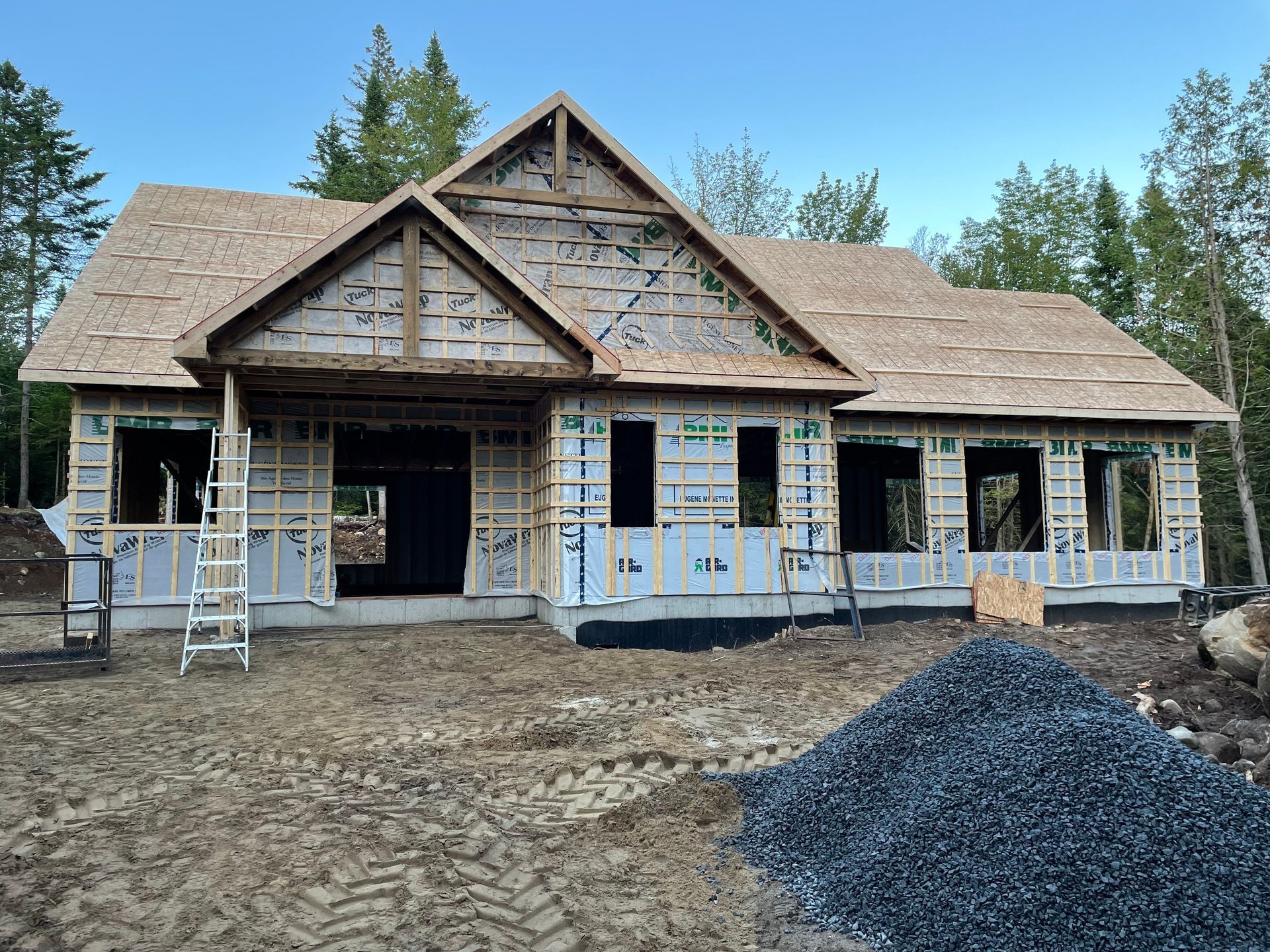 Maison en construction avec charpente apparente et toiture inachevée. Tas de gravier au premier plan.