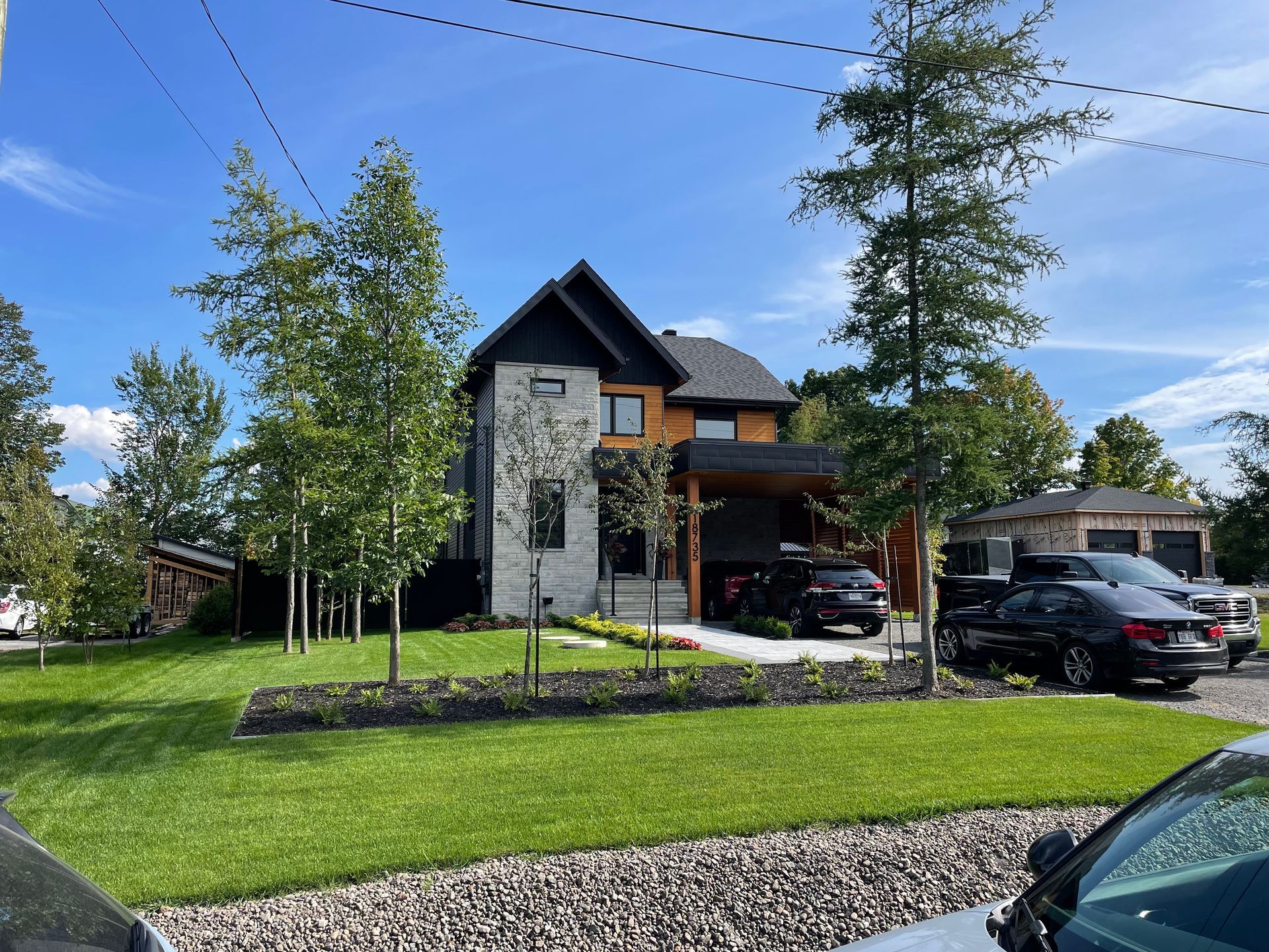 Maison moderne à deux étages avec façade en pierre, finitions noires et une voiture garée dans l'allée par une journée ensoleillée.